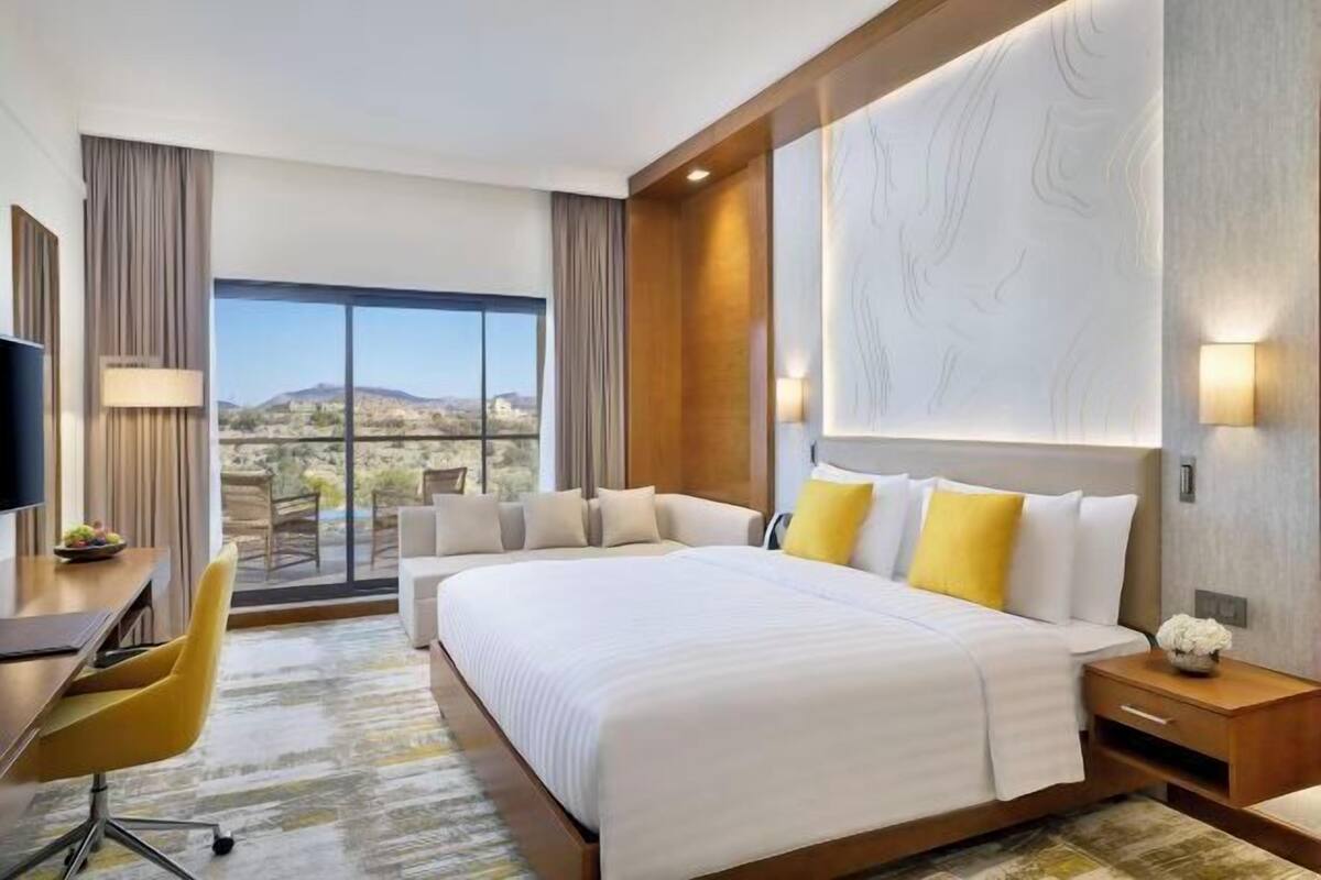 A hotel room with a large bed, a desk, and a chair. There is a window with a view of the mountains.