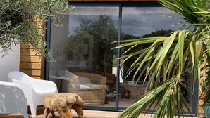 A modern outdoor patio with a glass door leading to a seating area, white plastic chairs, a wooden table, and a decorative bull sculpture. - Maison Avec Piscine (Saint-Jean-de-Monts)