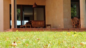 Un espacio de estar al aire libre cubierto con un banco y sillas de madera, una lámpara colgante y vista a un edificio con grandes ventanales.