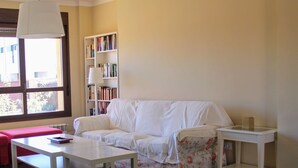 Una sala de estar con un sofá blanco, una mesita, una estantería y una ventana con persianas.