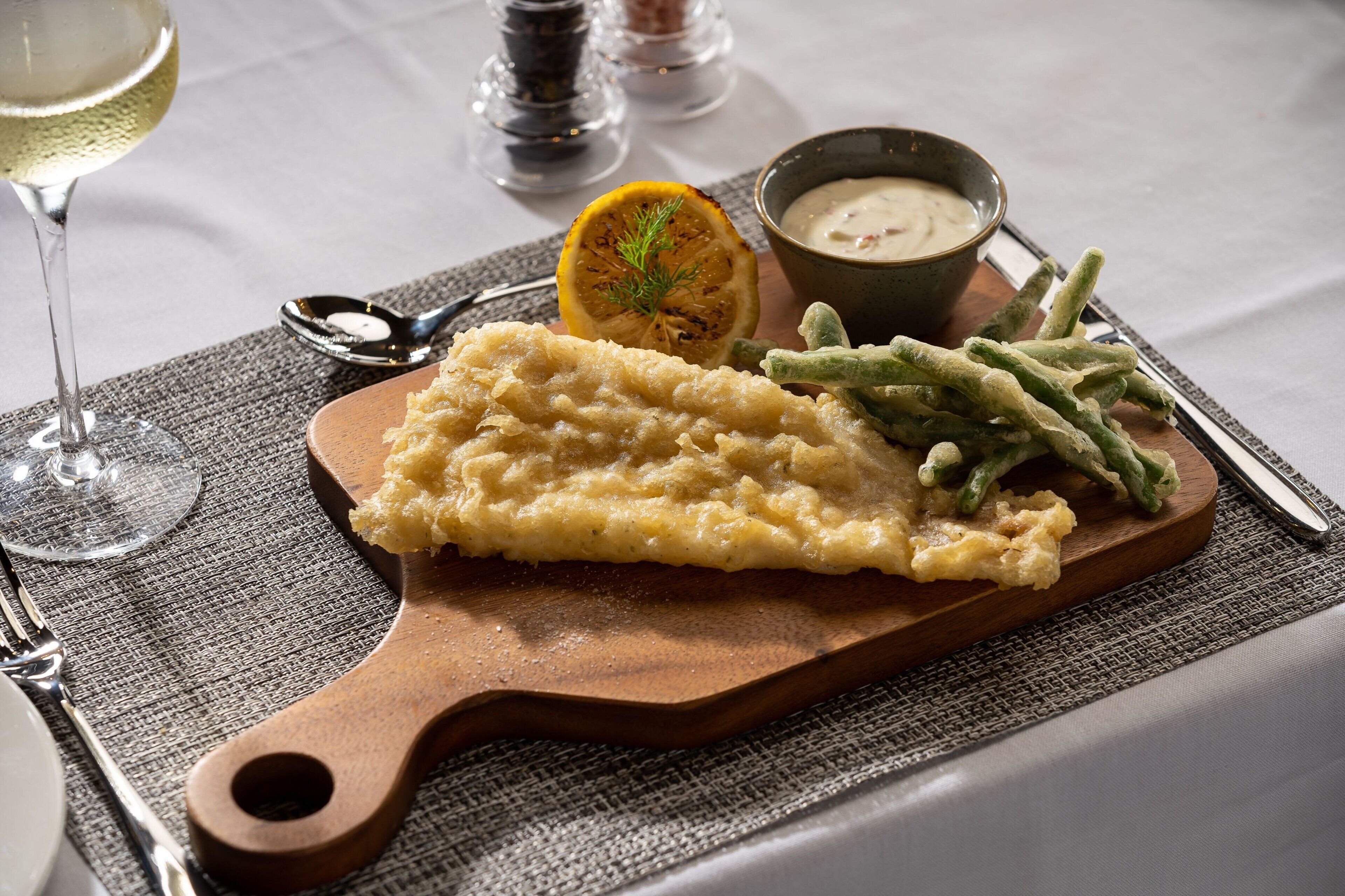 A wooden serving board with mashed potatoes, green beans, and a lemon slice on a tablecloth.
