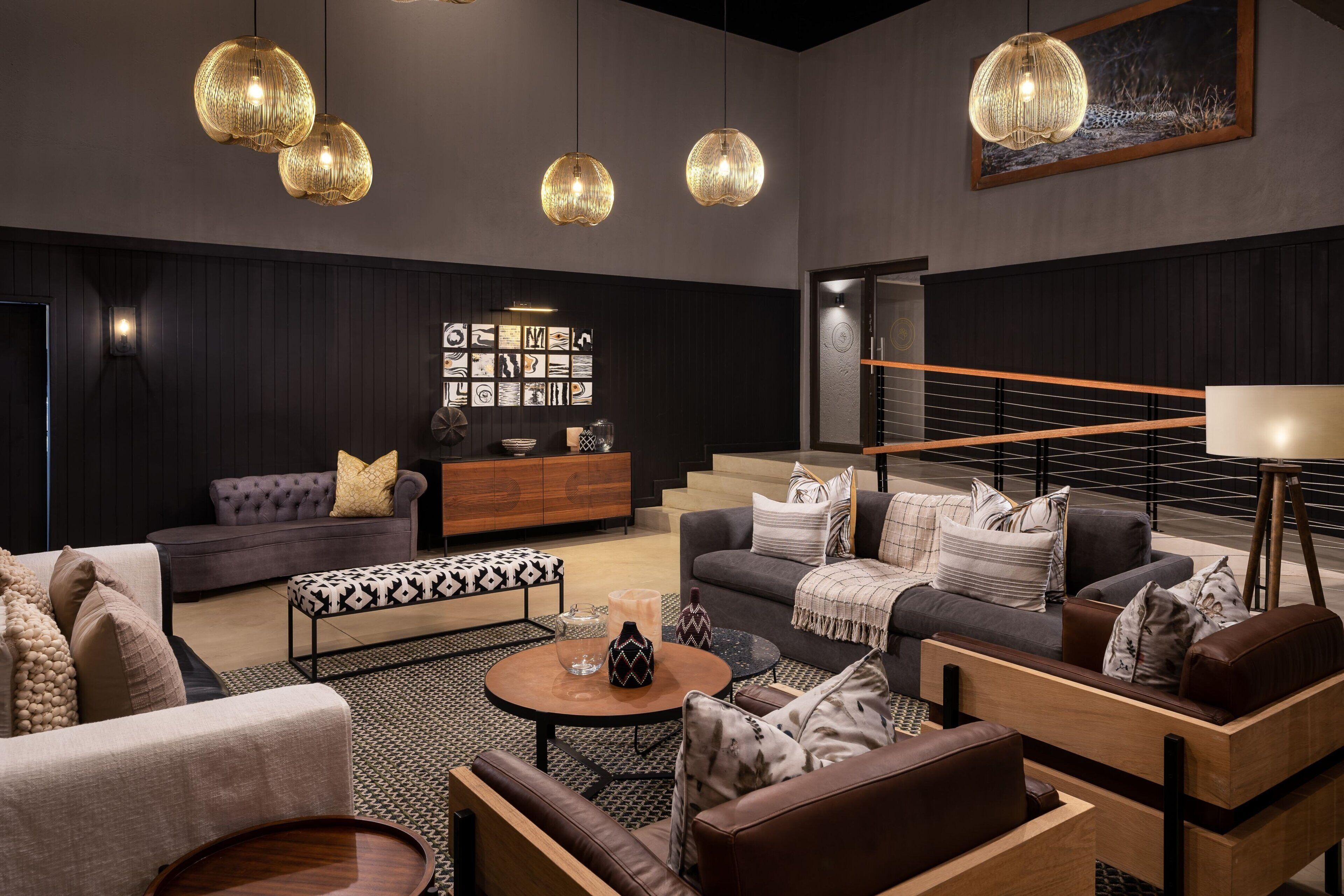 Una sala de estar moderna con paneles de madera oscura, un sofá gris, un reposapiés estampado y una mesa de centro. Se observan tres lámparas colgantes esféricas y una lámpara de pie grande.
