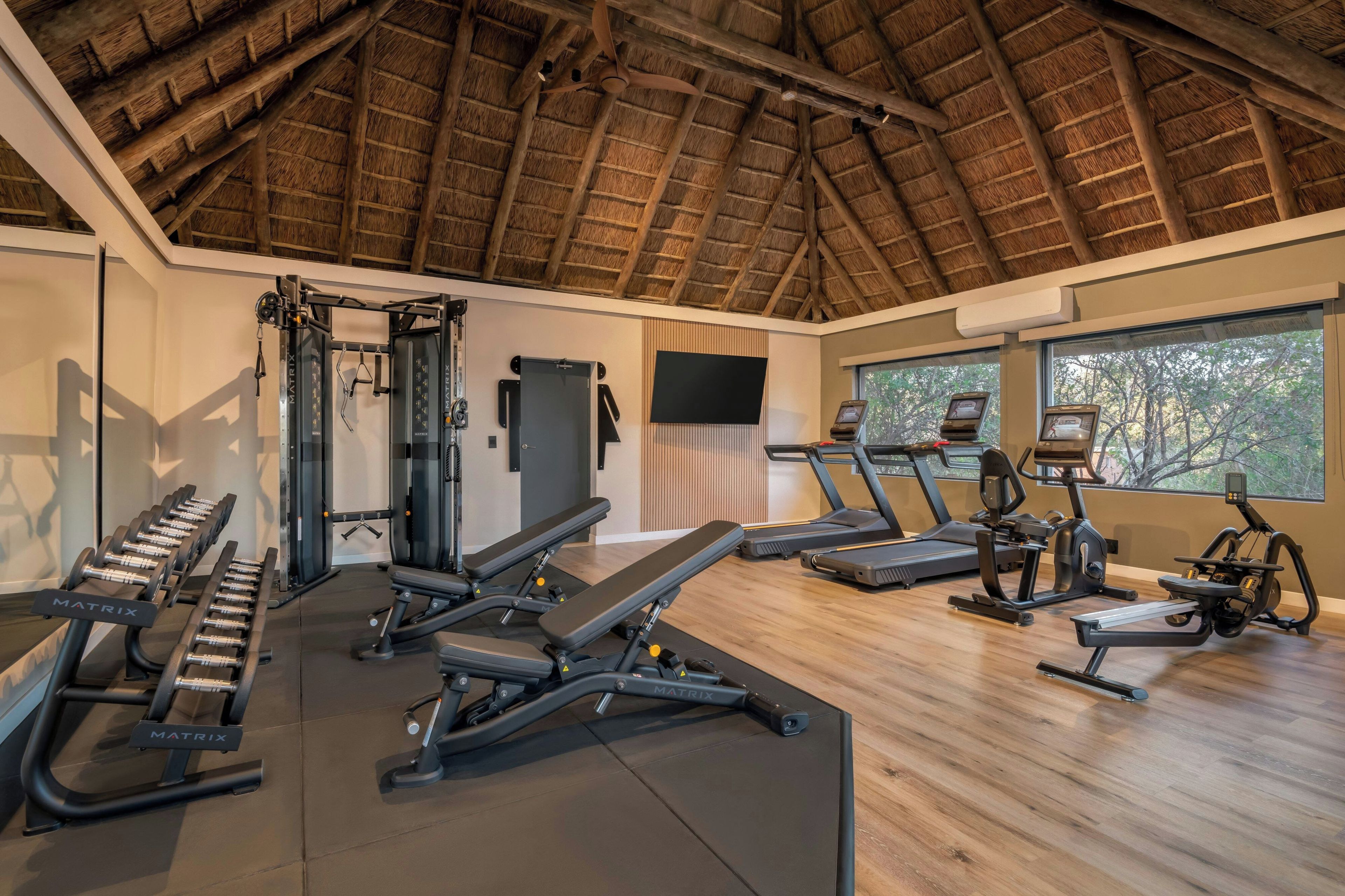 A gym with various exercise equipment, a large screen TV, and a thatched roof ceiling.