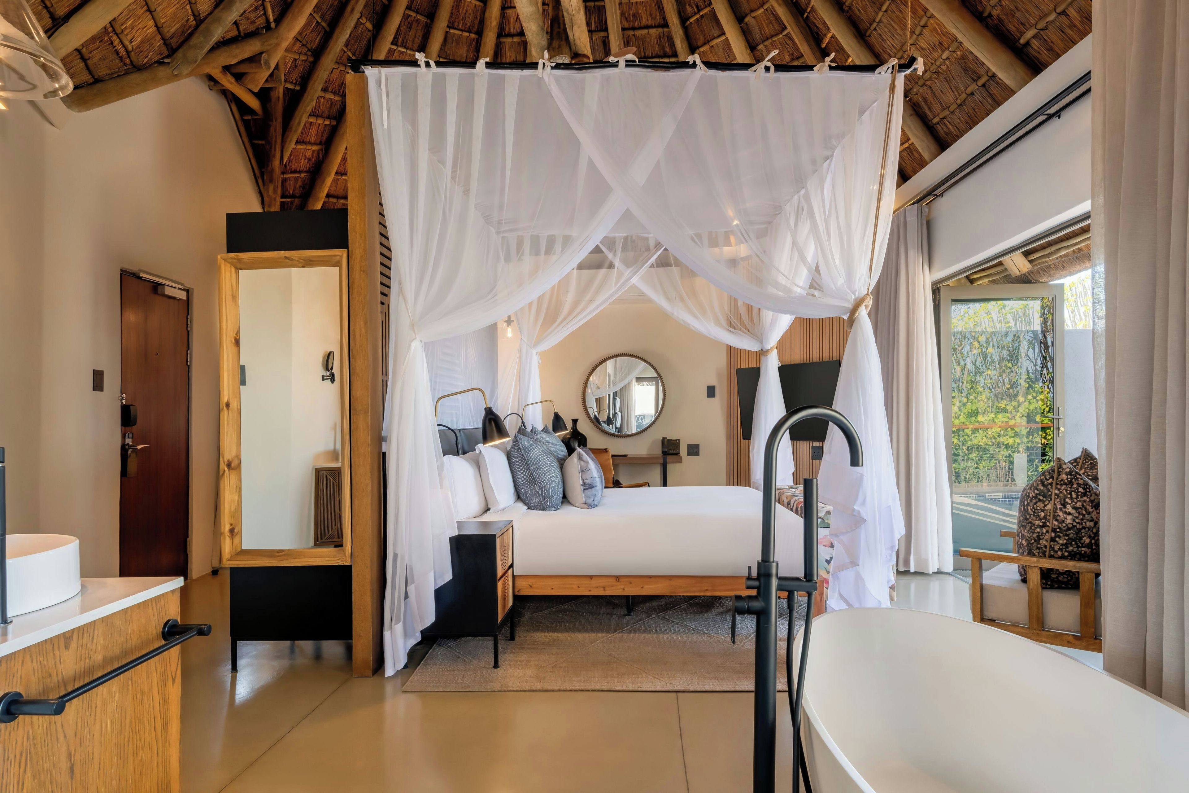 A bedroom with a canopy bed, a large mirror, bedside lamps, and a view of greenery outside.