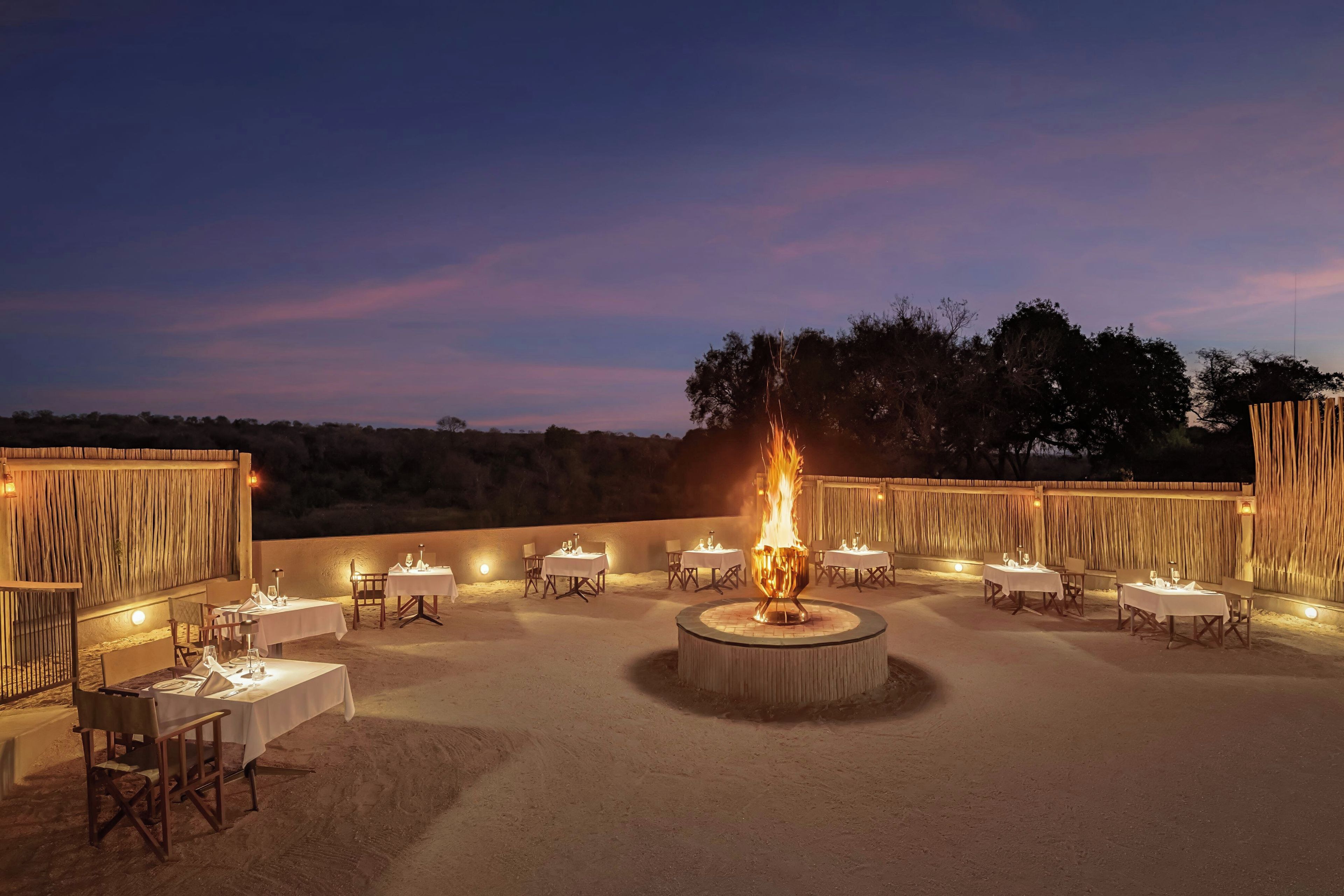 Un área de comedor al aire libre con mesas y sillas dispuestas alrededor de una chimenea central.