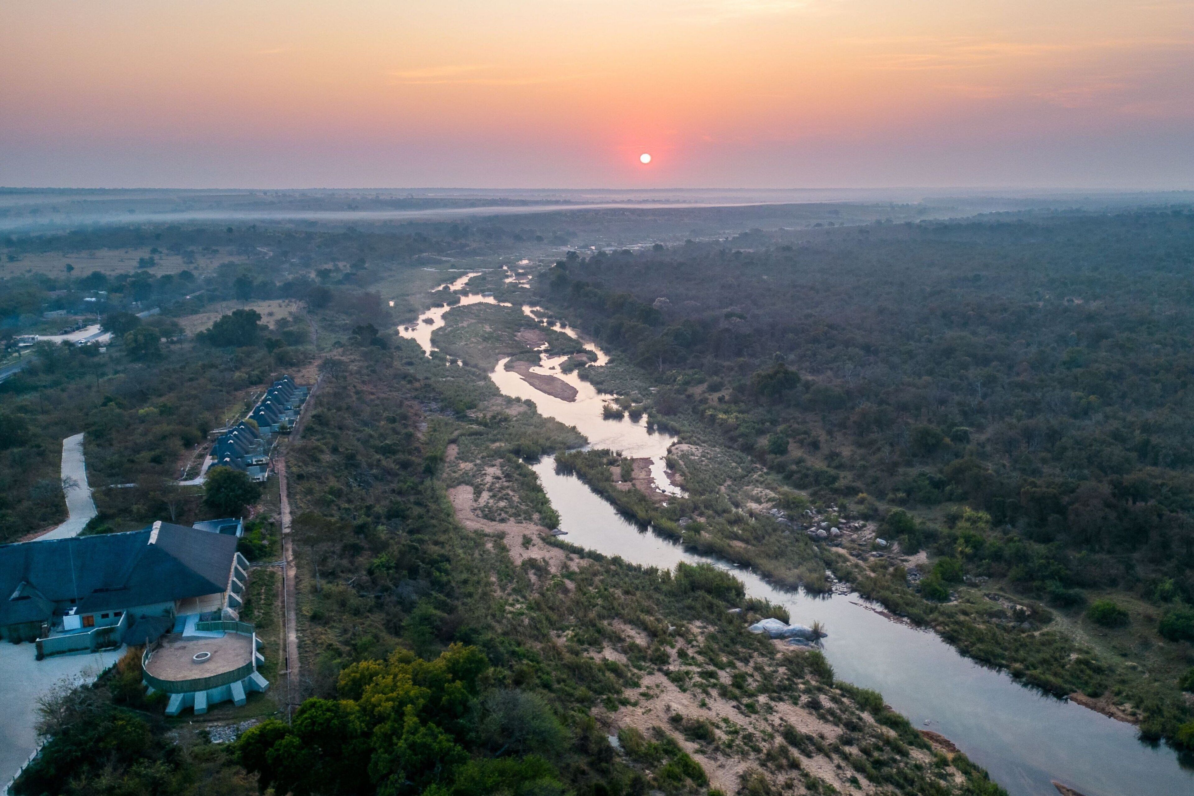 An aerial view of a river winding through a forested area with a building complex on the left.