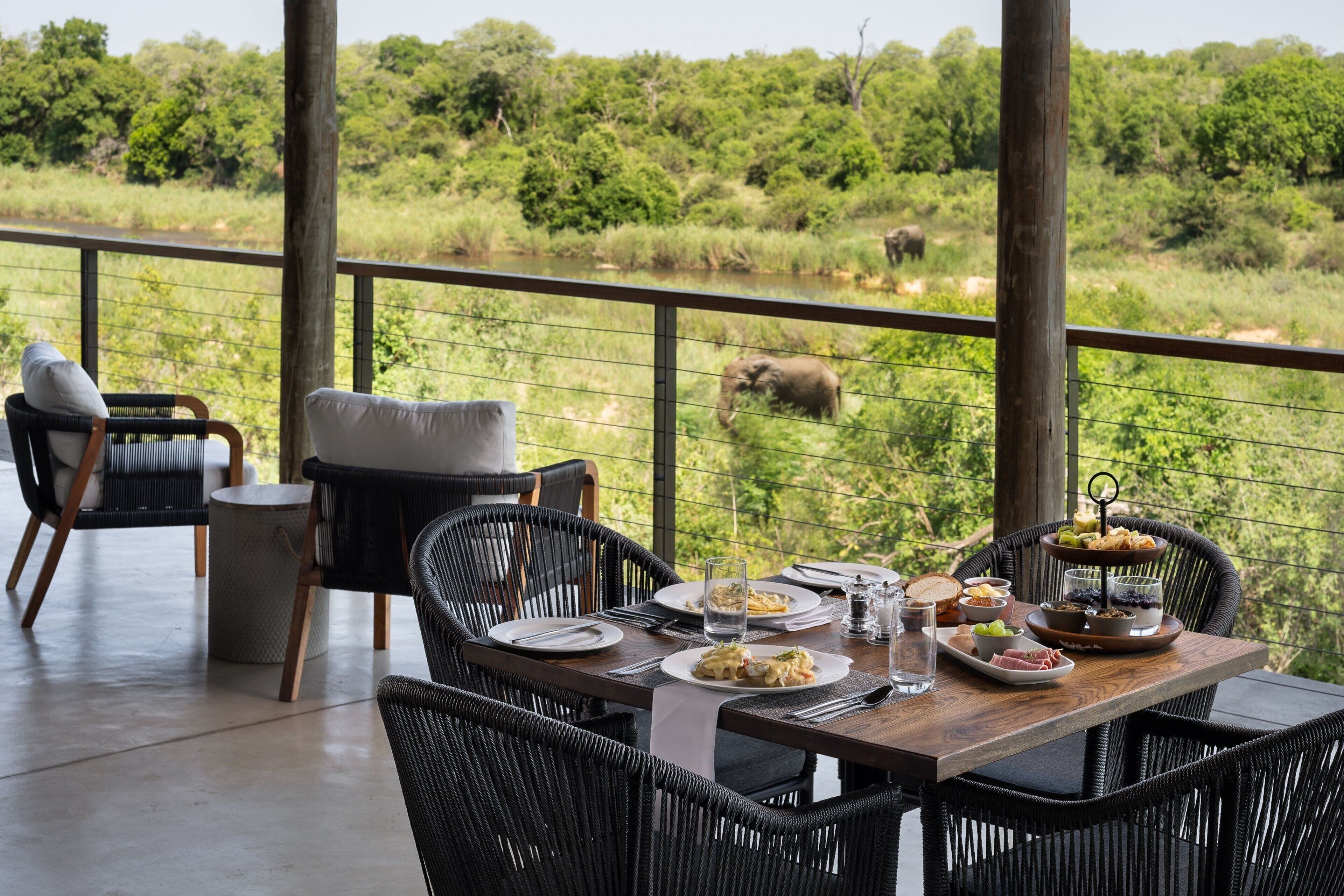 A dining area with wicker chairs and a wooden table set for a meal, overlooking a natural landscape with an elephant in the distance.