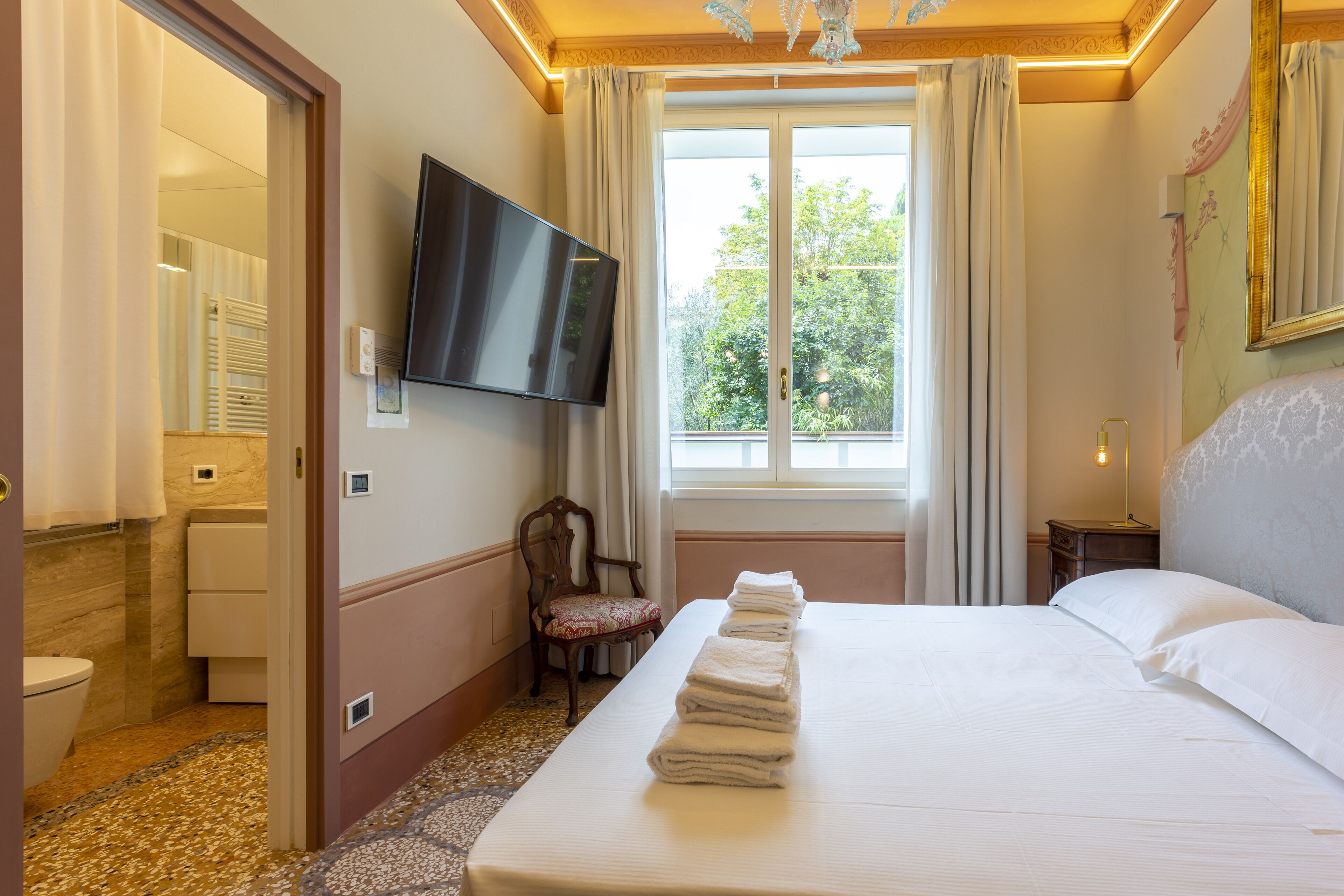 A hotel room with a bed, a TV mounted on the wall, a window with curtains, a chair, and a bathroom visible through an open door.
