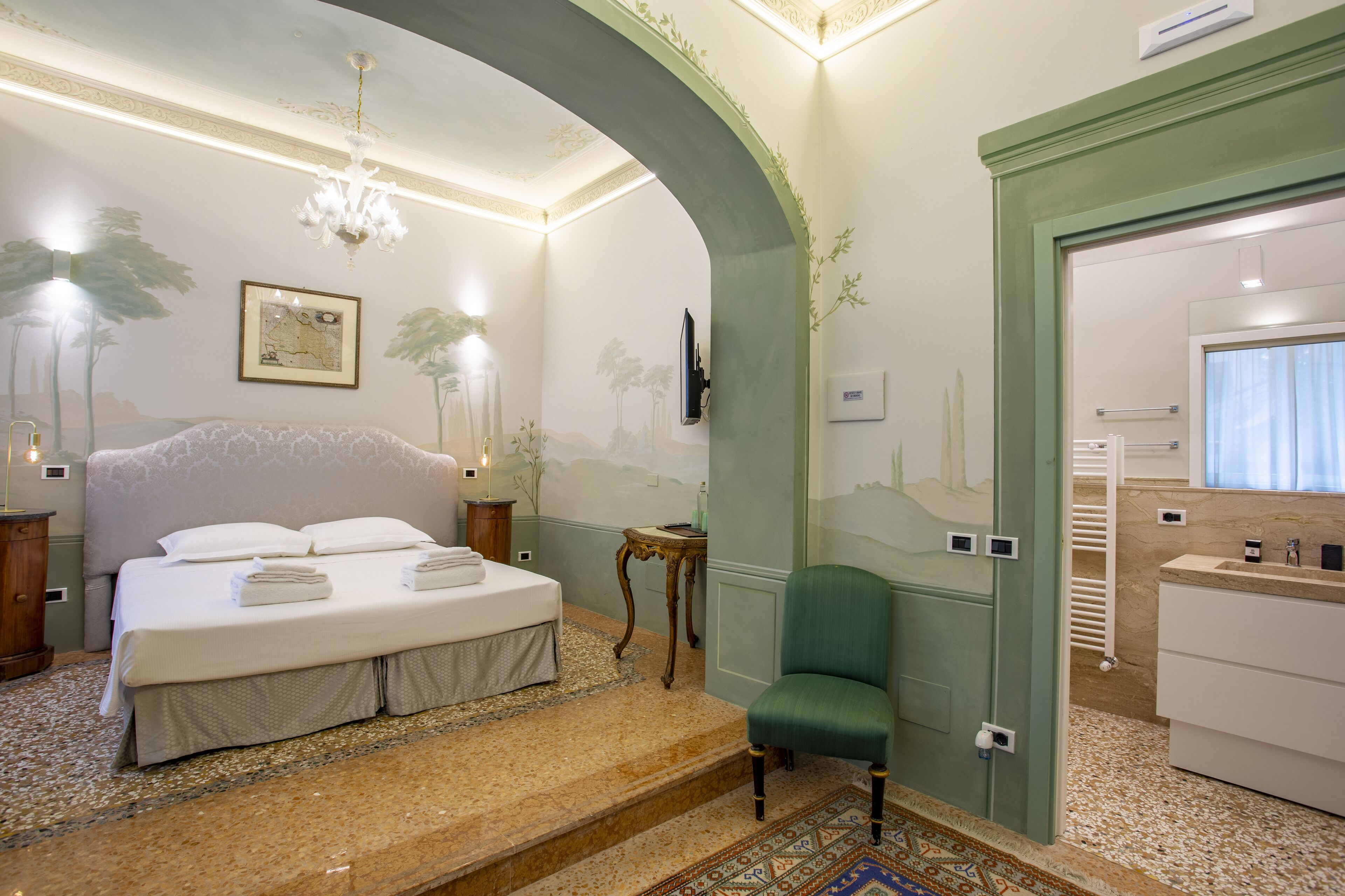 A bedroom with a large bed, a green armchair, a chandelier, and a bathroom visible through an archway.