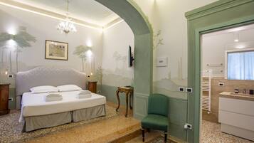 A bedroom with a large bed, a green armchair, a chandelier, and a bathroom visible through an archway.