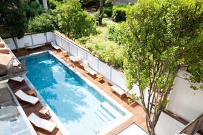 Piscina rectangular con cubierta de madera y sillas de descanso, rodeada de vegetación y una pared blanca.