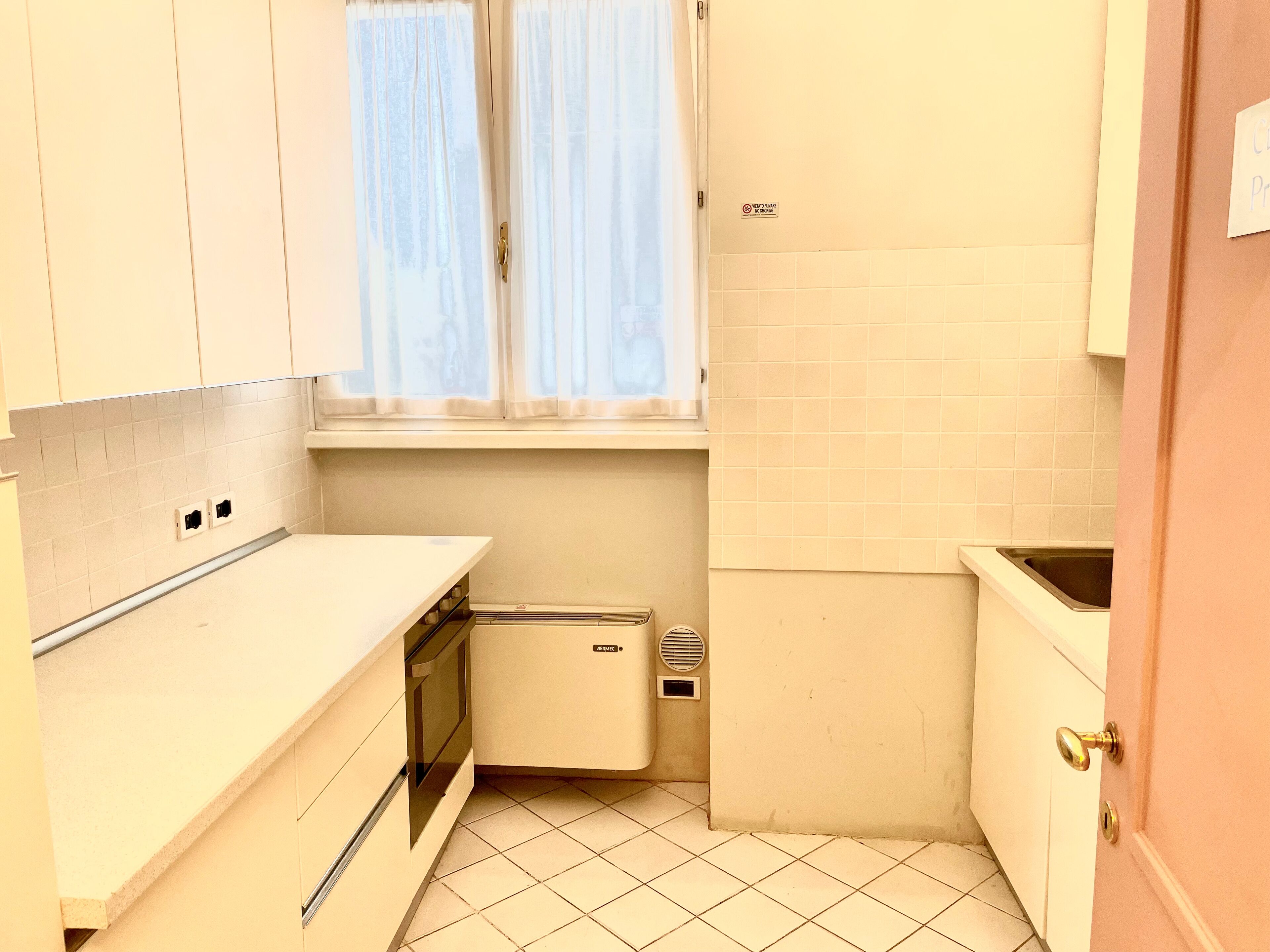 A compact kitchen with white cabinets, a small oven, a sink, and a window with curtains.