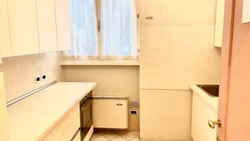 A compact kitchen with white cabinets, a small oven, a sink, and a window with curtains.