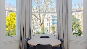 A room with a round table and chairs, large windows with curtains, and a view of buildings and trees. - Modern & Spacious 1 Bedroom Apartment in London (London)