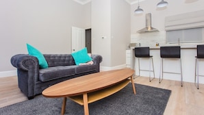 A modern living room with a grey sofa, teal cushions, a wooden coffee table, and a kitchen area with bar stools. - Modern & Spacious 1 Bedroom Apartment in London (London)