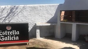 Placa de 'Estrella Galicia' e área de churrasco com parede branca e estruturas de concreto.