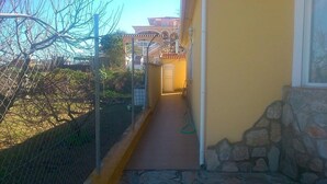 Um corredor estreito ao ar livre, com uma parede de pedra de um lado e uma grade de metal do outro, conduzindo a um edifício com telhado de cerâmica.