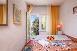 Una habitación de hotel con cama, mesita de noche, lámpara, ventana con cortinas y puerta que da a un balcón.