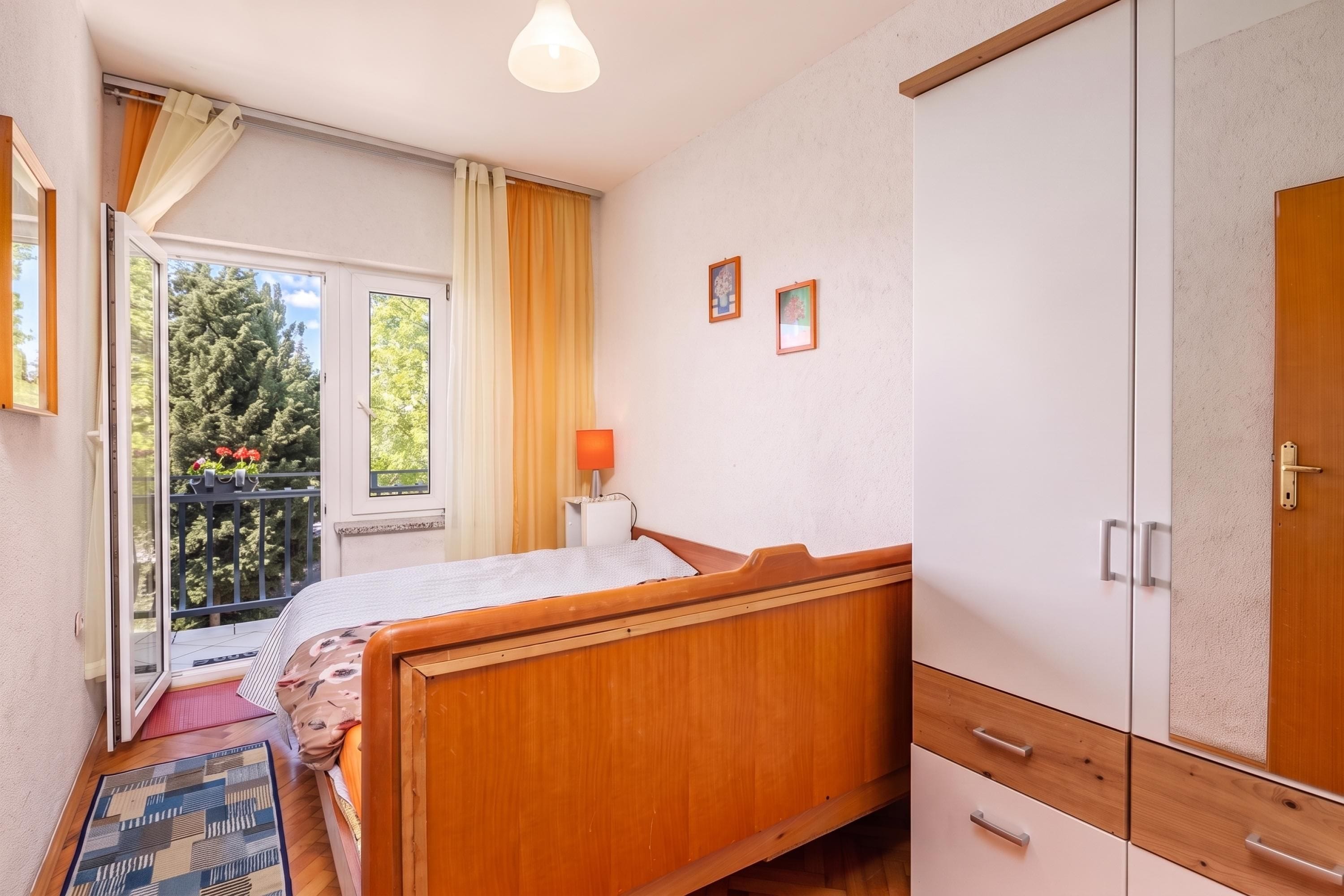 Un dormitorio con una cama de madera, un armario, una mesita de noche con una lámpara y una ventana con cortinas.