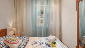 Camera d'albergo con un letto, un comodino con lampada, una finestra con tende leggere e vista esterna.