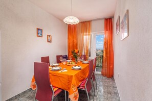 Uma sala de jantar com uma mesa posta para quatro pessoas, cadeiras, um lustre e vista por uma porta.