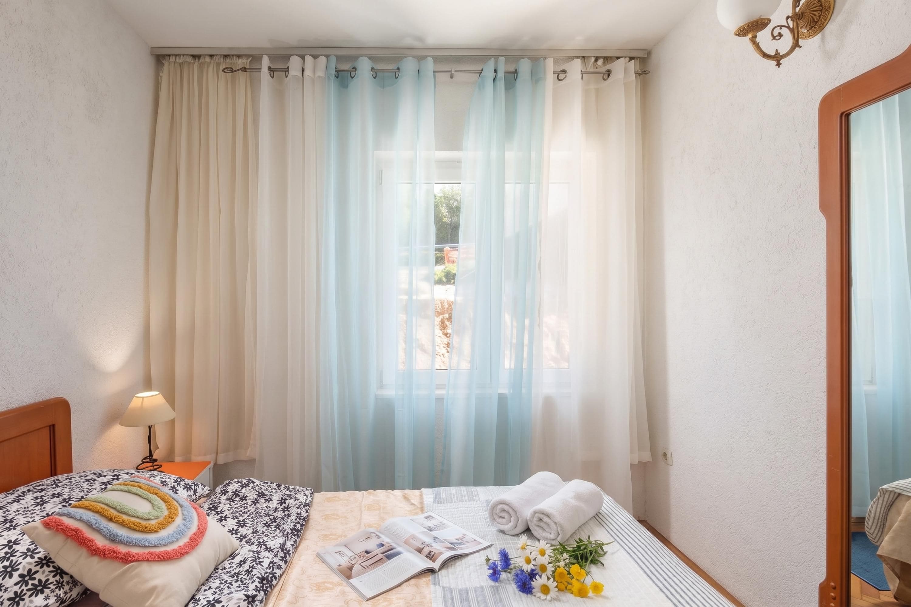 A hotel room with a bed, a nightstand, a lamp, a window with sheer curtains, and a view of the outside.