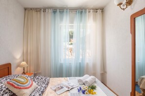 A hotel room with a bed, a nightstand, a lamp, a window with sheer curtains, and a view of the outside. - Seaview Apartment Micika Krk Island (Krk)