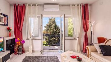 Ein modernes Wohnzimmer mit einer Couch, einem Couchtisch und einem Fernseher. Es gibt einen Balkon mit Blick auf Bäume und einen Berg.