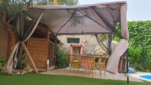 A covered outdoor seating area with a table and chairs, surrounded by a garden and a pool. - Dependence 2 Fronte Piscina in Centro Storico (Oristano)