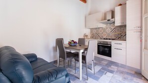 A compact living space with a sofa, dining table, chairs, and a kitchen area including cabinets, a stove, and an oven. - Dependence 2 Fronte Piscina in Centro Storico (Oristano)