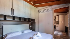 A hotel room with a large bed, white linens, and neatly folded towels. There is a wooden ceiling and a kitchenette in the background. - Dependence 2 Fronte Piscina in Centro Storico (Oristano)