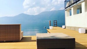 Una terraza de madera con dos sillones de mimbre y una mesita, con vistas a un cuerpo de agua y montañas.