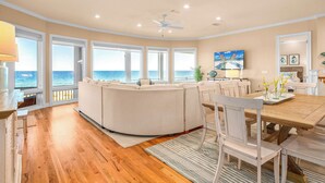 A spacious living area with a curved sofa, a dining table with chairs, and large windows offering a view of the ocean. - CJ's Beach Front 6 BR Heated Pool Private Home (St. George Island)