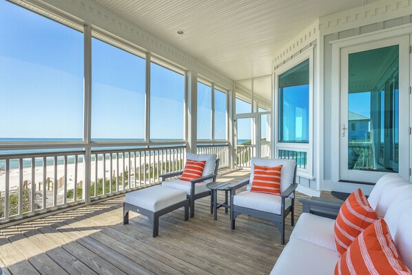 Un espace extérieur abrité doté d’une terrasse en bois, de mobilier blanc et de grandes fenêtres offrant une vue sur la plage.