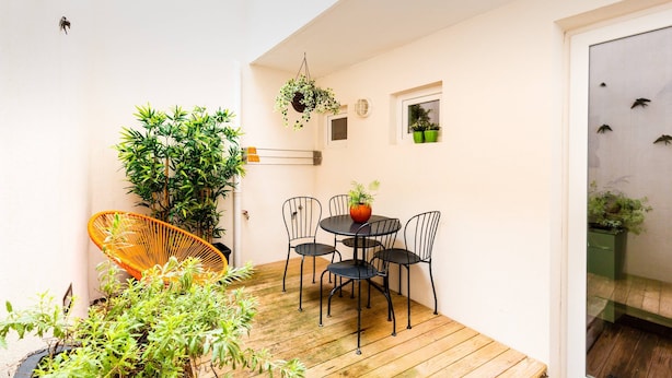 Un patio con una terraza de madera, una mesa redonda con cuatro sillas, una planta colgante y una silla de mimbre.