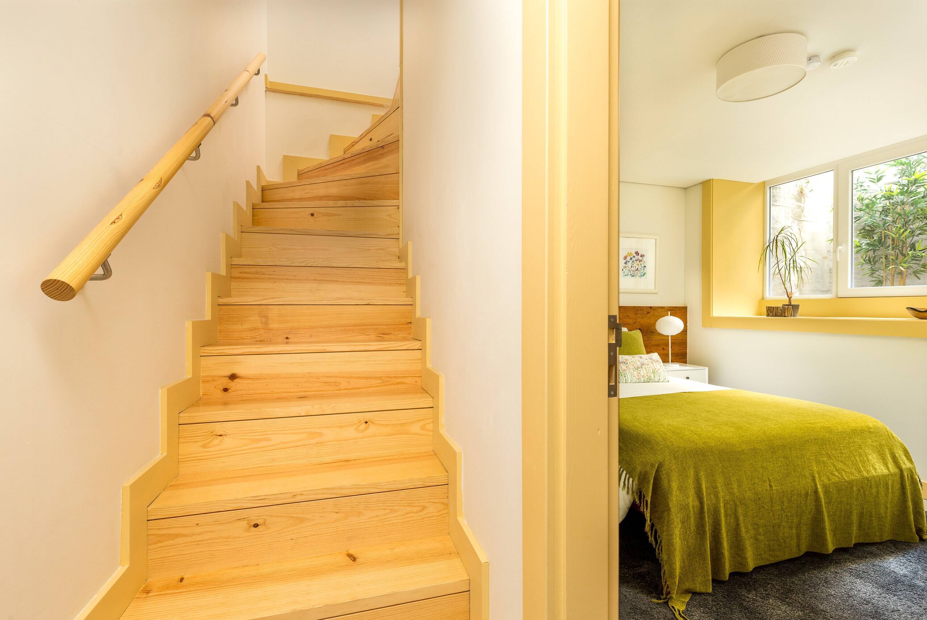 Una escalera de madera con barandilla que lleva a una habitación con cama, ventana y mesita de noche.