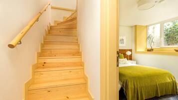 Una escalera de madera con barandilla que lleva a una habitación con cama, ventana y mesita de noche.