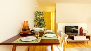 Comedor con una mesa de madera dispuesta para cuatro personas, un televisor en un mueble bajo y una planta en maceta al fondo.