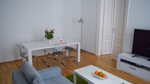 A modern living room with a grey sofa, a white dining table with chairs, a small table with fruit and a remote, a TV on a white stand, and a floor lamp. - Modern apartment in Vienna near Danube (Vienna)