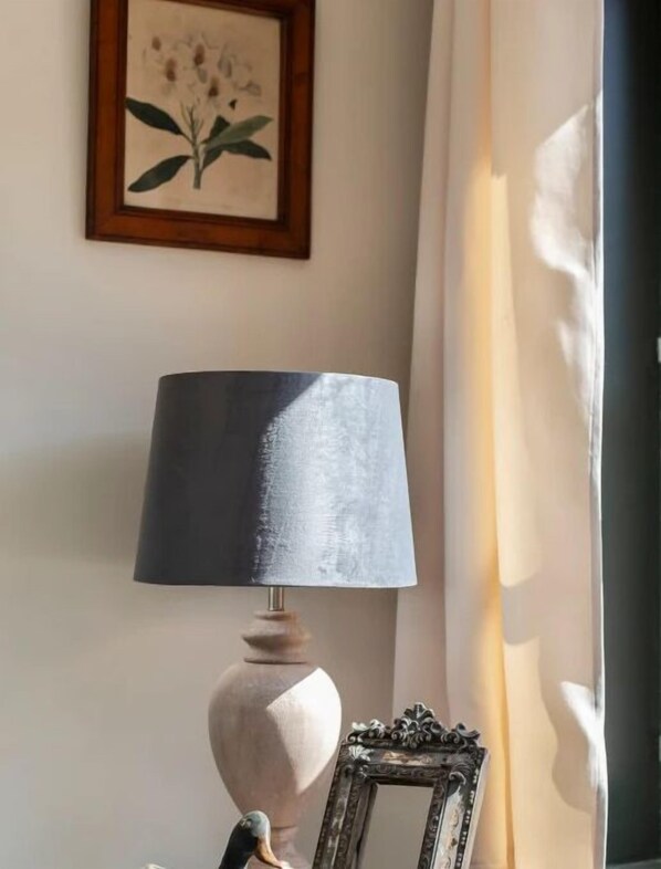 Eine Lampe mit dunklem Schirm, ein gerahmtes Bild an der Wand und ein zur Seite zurückgezogener Vorhang.