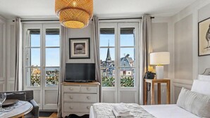 Une chambre d’hôtel équipée d’un lit, d’une télévision, d’une lampe, d’une table à manger et offrant une vue sur le paysage urbain grâce à de grandes fenêtres.