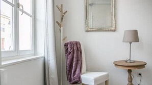 Una habitación con una silla blanca, una mesa redonda con una lámpara, un espejo en la pared y una ventana con cortinas.
