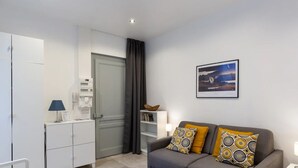 Uma sala moderna com um sofá cinzento, duas almofadas estampadas, um quadro na parede, um armário branco e uma porta cinzenta.