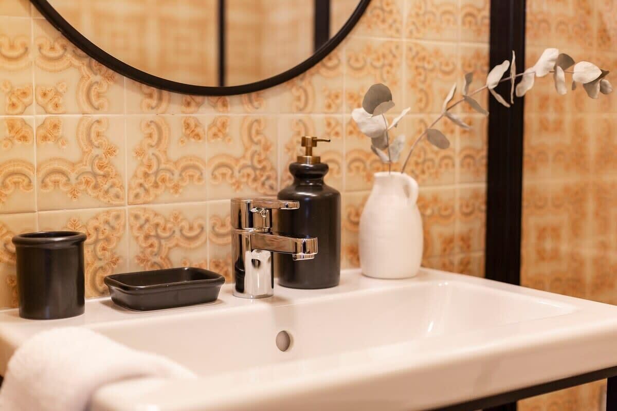 Uma pia de banheiro com espelho, um dosador de sabonete, um vaso com um galho e um porta-objetos preto.