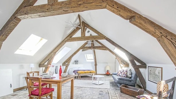 Camera mansardata con tetto spiovente, travi in legno, un letto, un divano e una zona pranzo con tavolo e sedie.