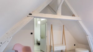 Une pièce avec un plafond en pente, un mur peint en blanc, une échelle en bois, un petit meuble blanc et une chaise rose.