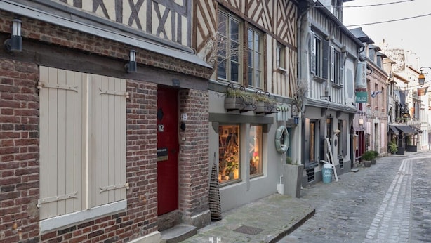 Une ruelle étroite pavée, bordée de bâtiments traditionnels à colombages, dont certains arborent des devantures de boutiques et des enseignes bien visibles.