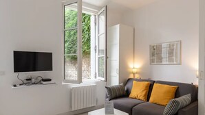 Un salon moderne doté d’un téléviseur à écran plat, d’un canapé avec des coussins, d’une fenêtre avec une porte ouverte et d’un luminaire fixé au mur.