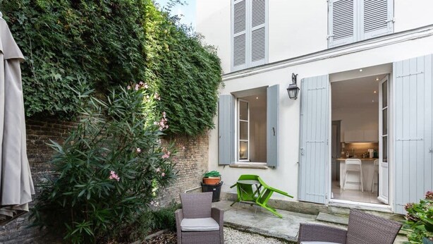 Une cour avec une chaise en osier, une chaise verte et un parasol beige. On y trouve un mur en briques et une plante aux fleurs roses.