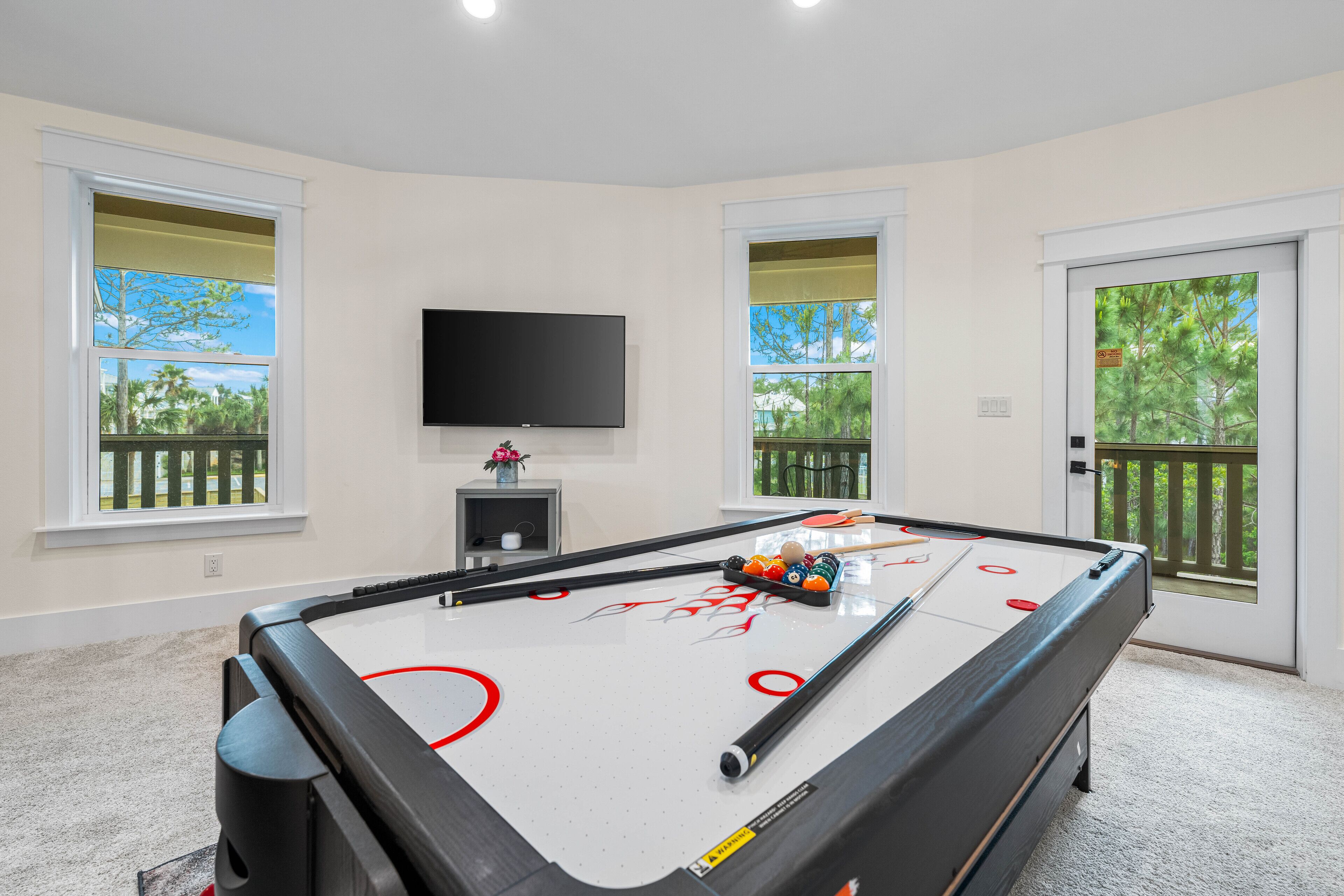 A pool table with a mounted TV, windows with views of trees, and a door leading to an outdoor deck.
